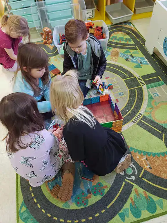 Preschool students at Lewis County Adventist School engaged in collaborative play activities on a colorful classroom rug, with a teacher guiding small group learning